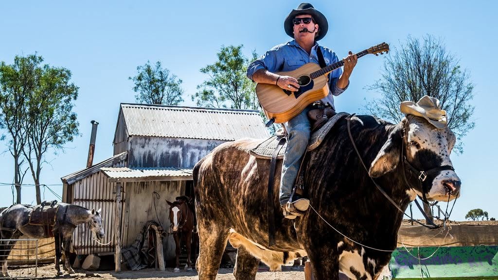Lachie Cossor playing guitar while riding his big bull past an old tin shed